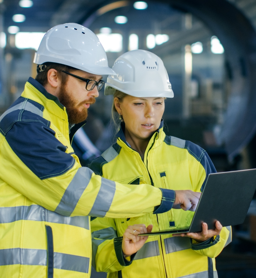 Bild von einem Mann mit Bart und eine Frau, beide in gelben reflektierenden Sicherheitsjacken und weißen Schutzhelmen. Sie stehen in einer Industriehalle. Der Mann zeigt auf den Bildschirm eines Laptops, den die Frau hält. Beide wirken konzentriert, während sie technische Details besprechen.