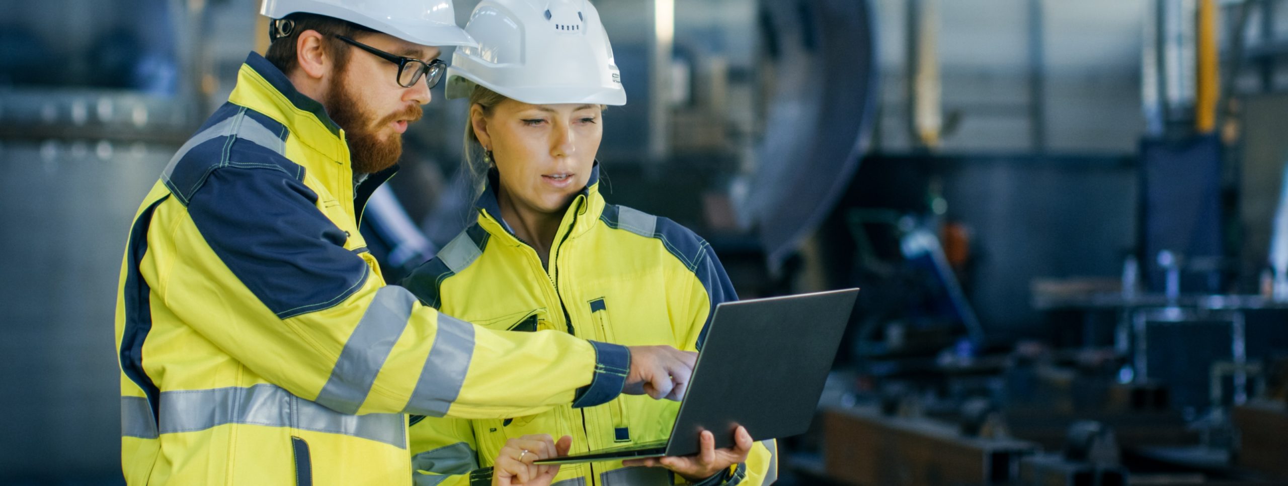 Bild von einem Mann mit Bart und eine Frau, beide in gelben reflektierenden Sicherheitsjacken und weißen Schutzhelmen. Sie stehen in einer Industriehalle. Der Mann zeigt auf den Bildschirm eines Laptops, den die Frau hält. Beide wirken konzentriert, während sie technische Details besprechen.